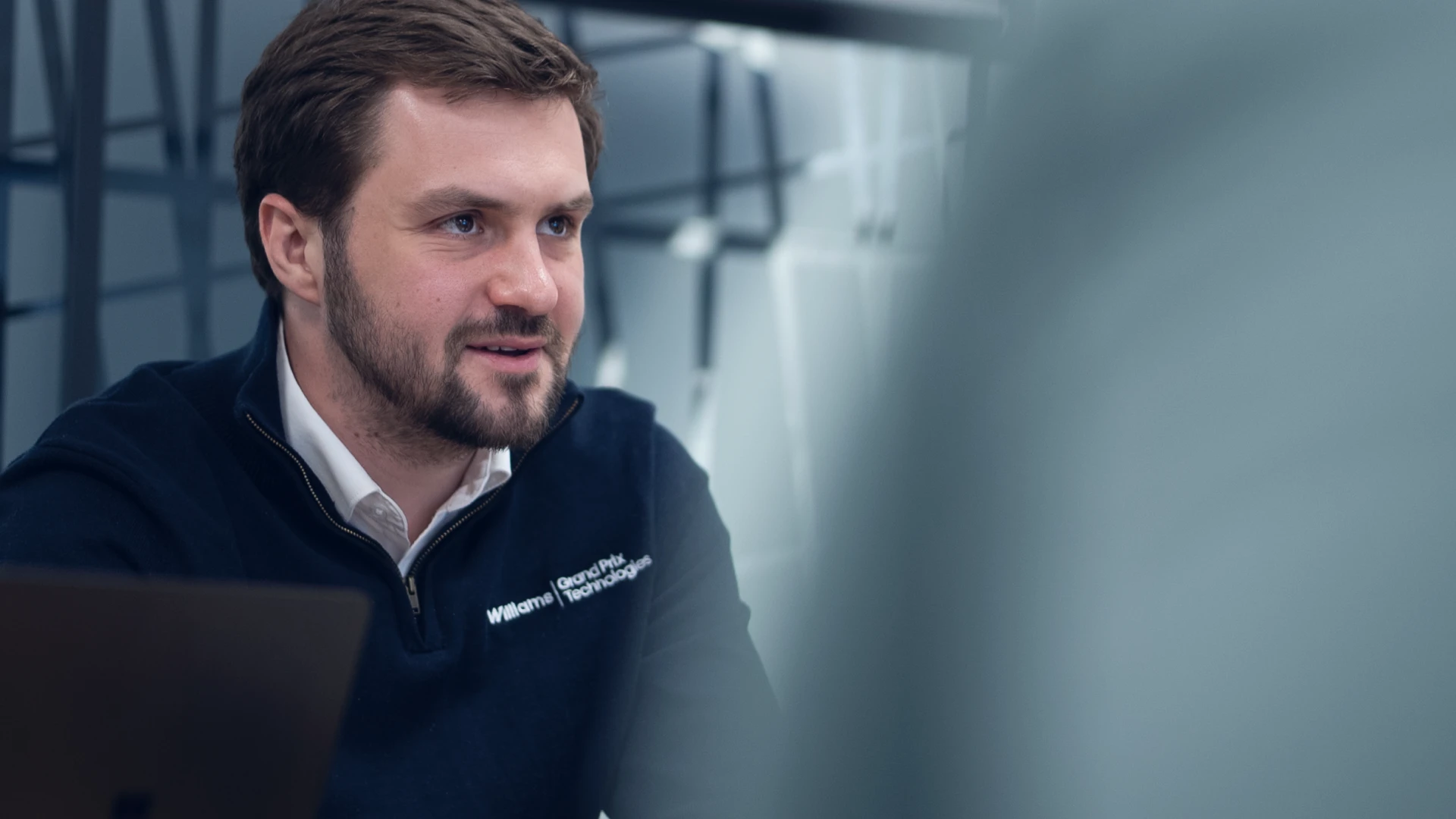 Man wearing Williams Grand Prix Technologies sweater engaged in conversation indoors.