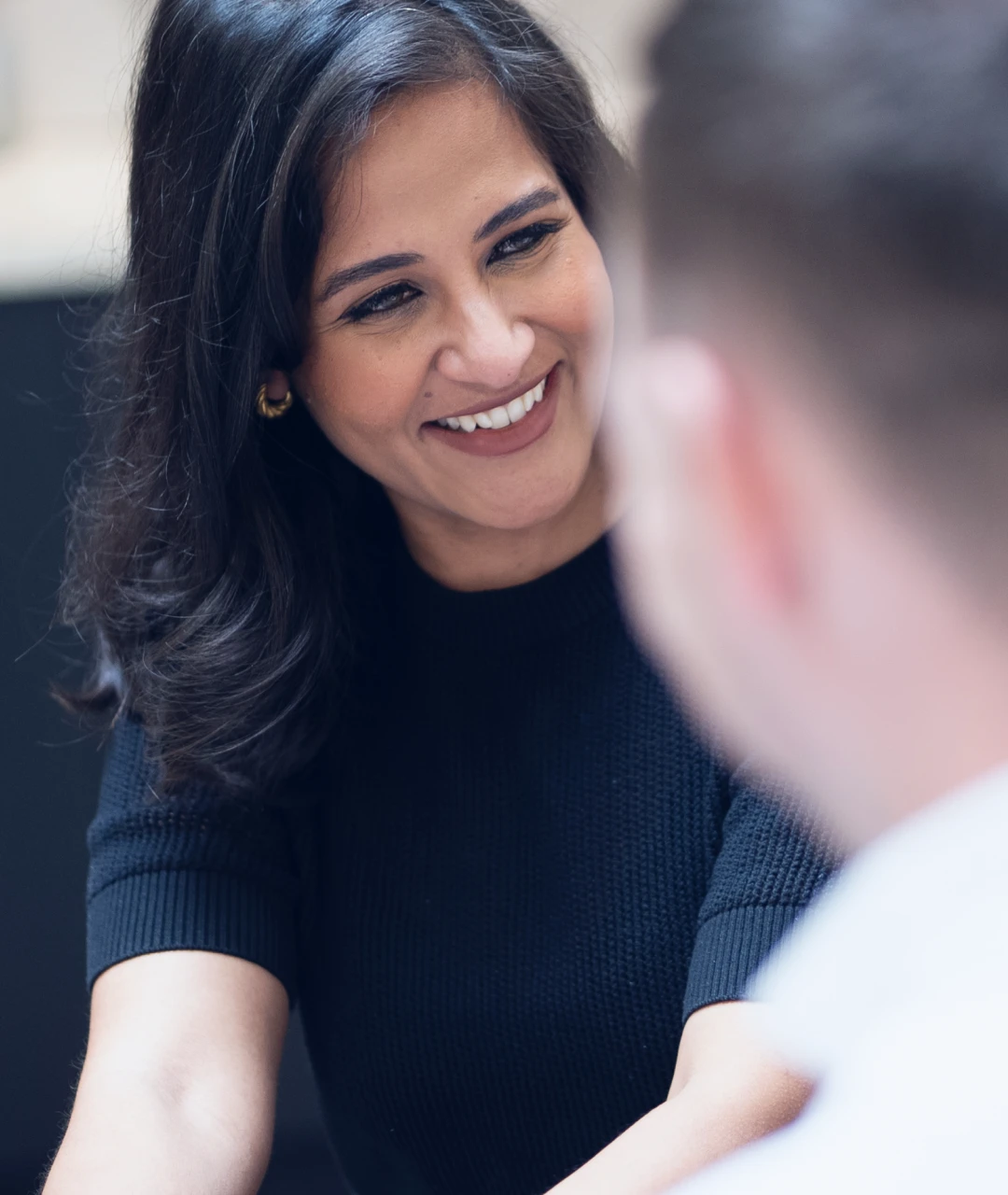 Smiling woman with dark hair wearing a black sweater engaging with a man in a white shirt.