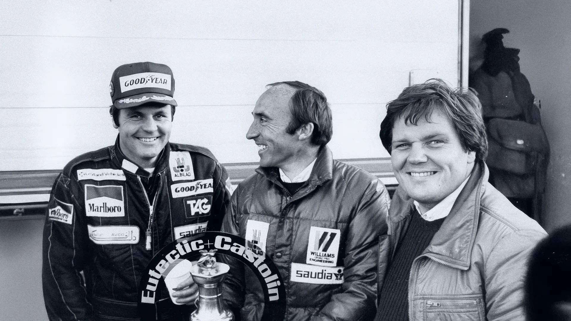 Three men smiling, two wearing racing suits with sponsor logos, holding a trophy ring labeled Eutectic+Castolin.