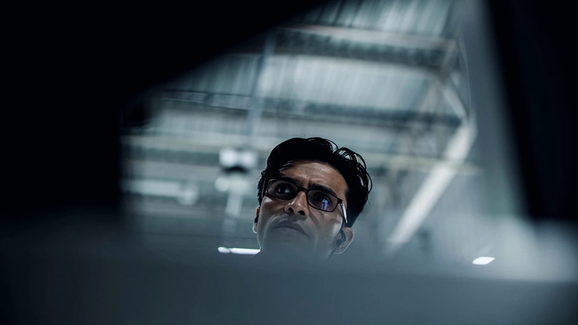 Focused man with glasses and earbuds looking upward in a dimly lit industrial setting.