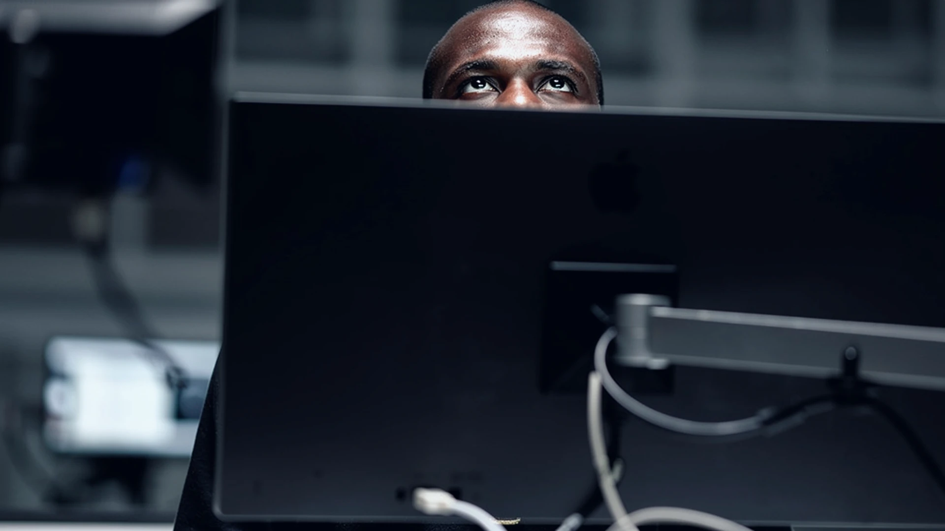 Man's eyes looking over the top edge of a large Apple computer monitor in a dim office.