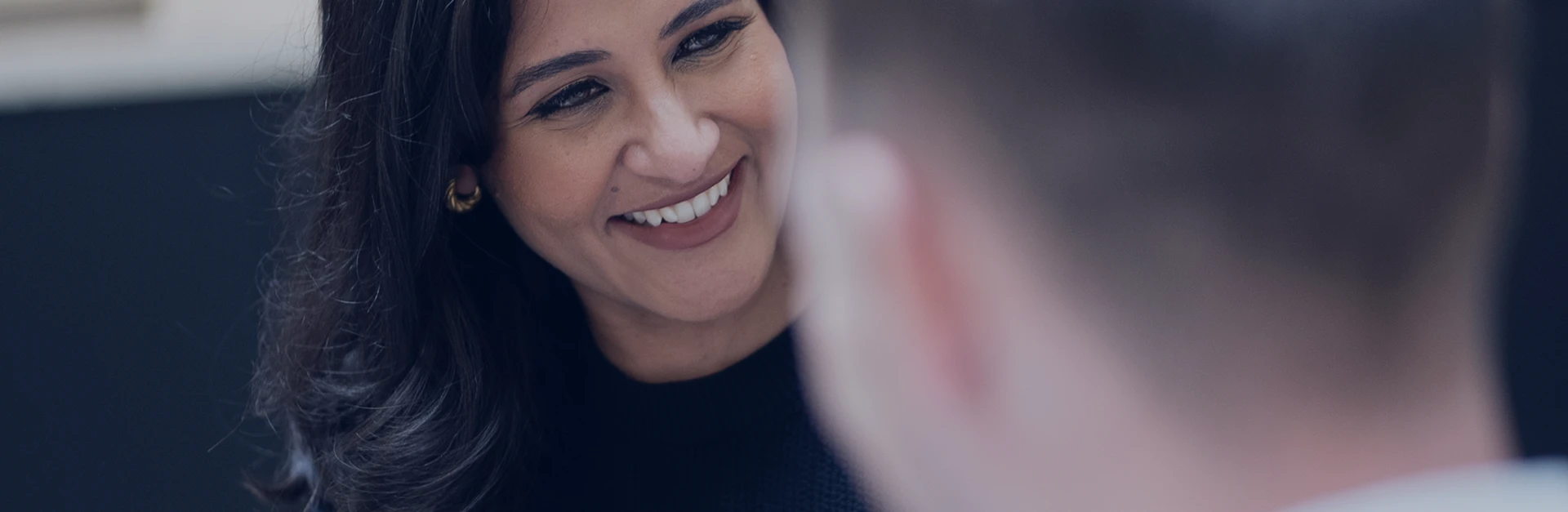 Smiling woman with long hair wearing gold earrings talking to a person with short hair.