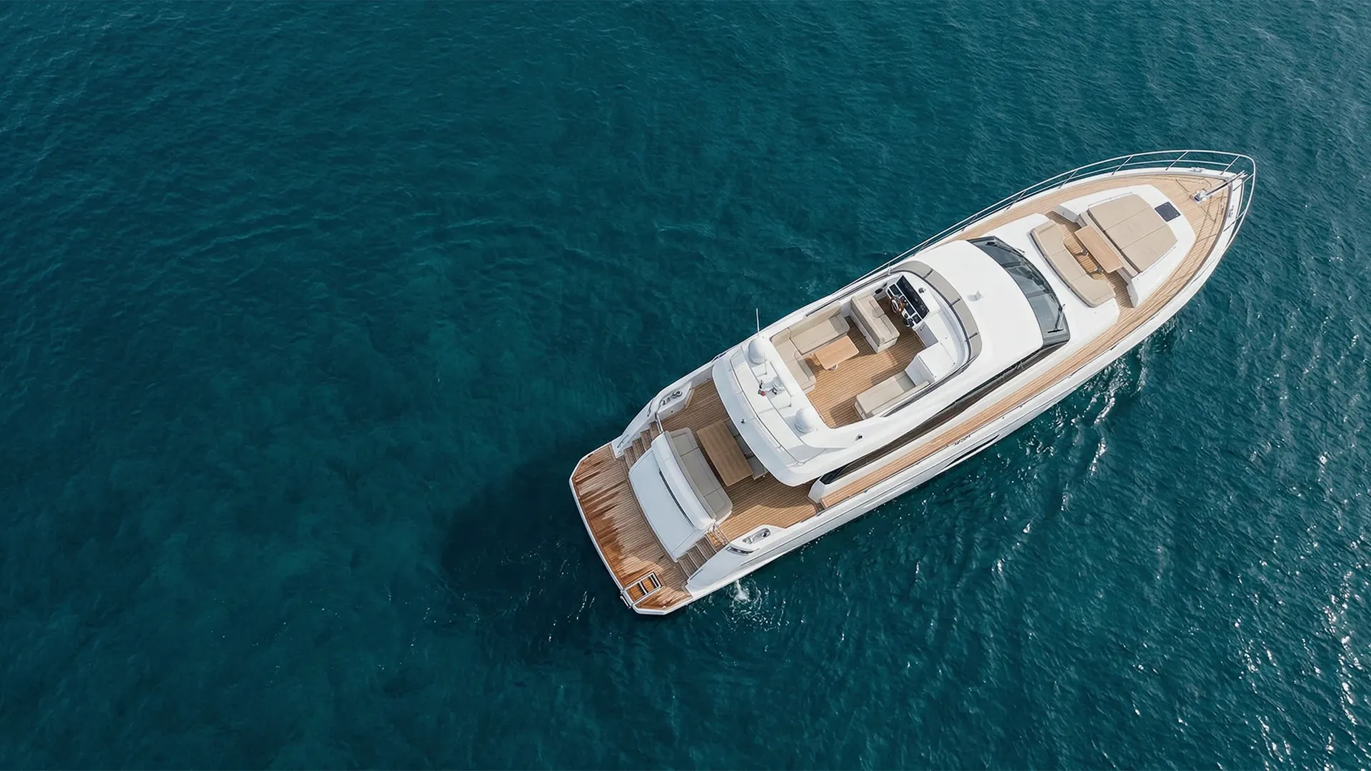 A white motor yacht with teak decks floating on clear deep blue water, viewed from above.