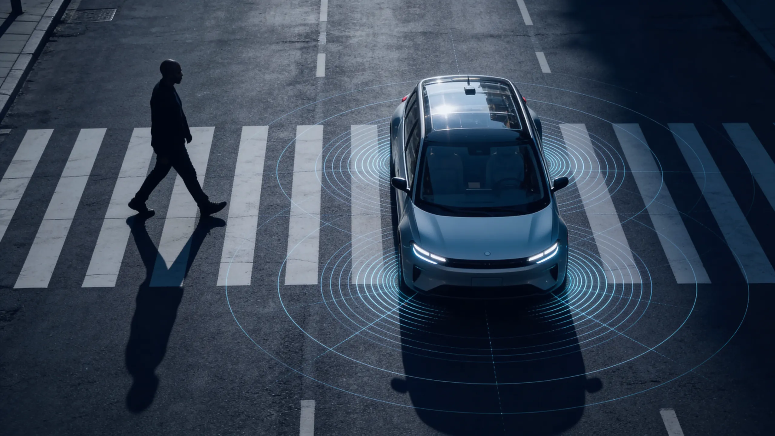 Self-driving car stops at crosswalk as pedestrian walks across the street in dark lighting.