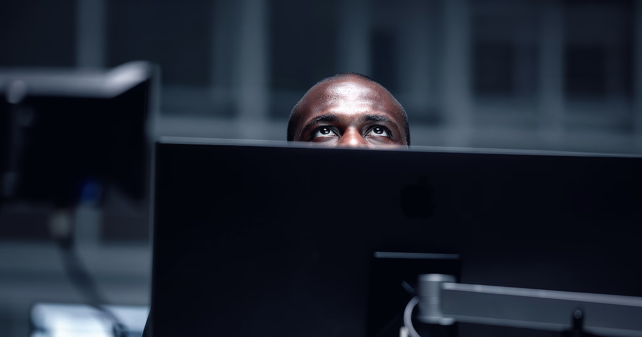 Man's eyes looking over the top edge of a large Apple computer monitor in a dim office.