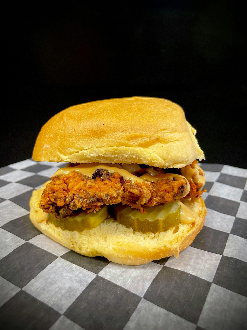 Fried chicken sandwich with pickles and sauce on a soft bun placed on black and white checkered paper.