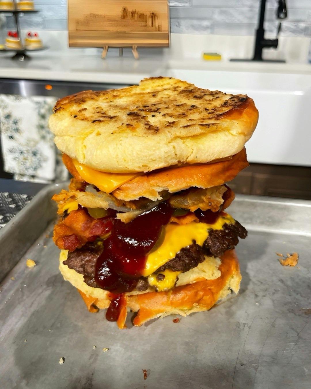 Thick cheeseburger with bacon, fried onions, melted cheddar, barbecue sauce, and an English muffin bun on a metal tray.