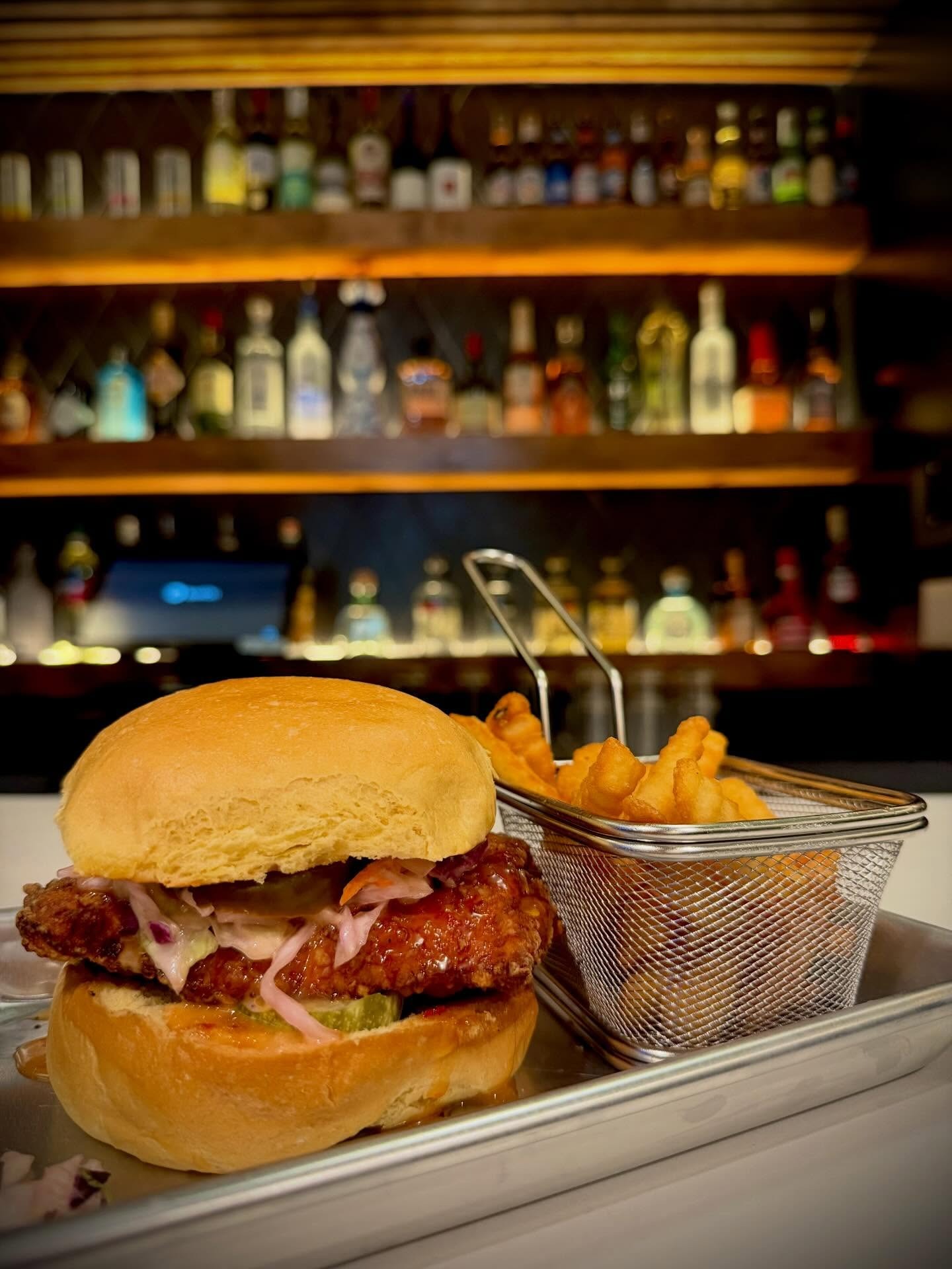 Spicy fried chicken sandwich with pickles and slaw on a bun served with crinkle-cut fries in a small metal basket, set against a blurred background of liquor bottles on shelves.