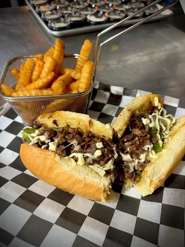 Philly cheesesteak sandwich with melted cheese and green peppers served with crinkle-cut fries in a metal basket on a black and white checkered paper.