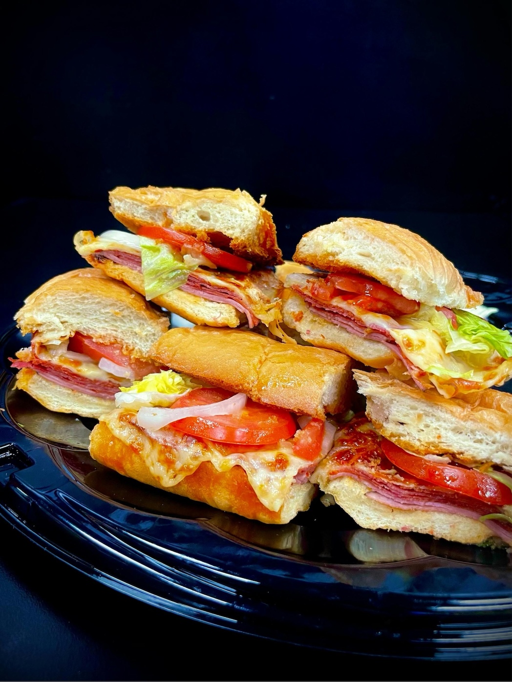 Plate with several sandwiches containing sliced meat, melted cheese, tomato, lettuce, and onions on bread rolls.