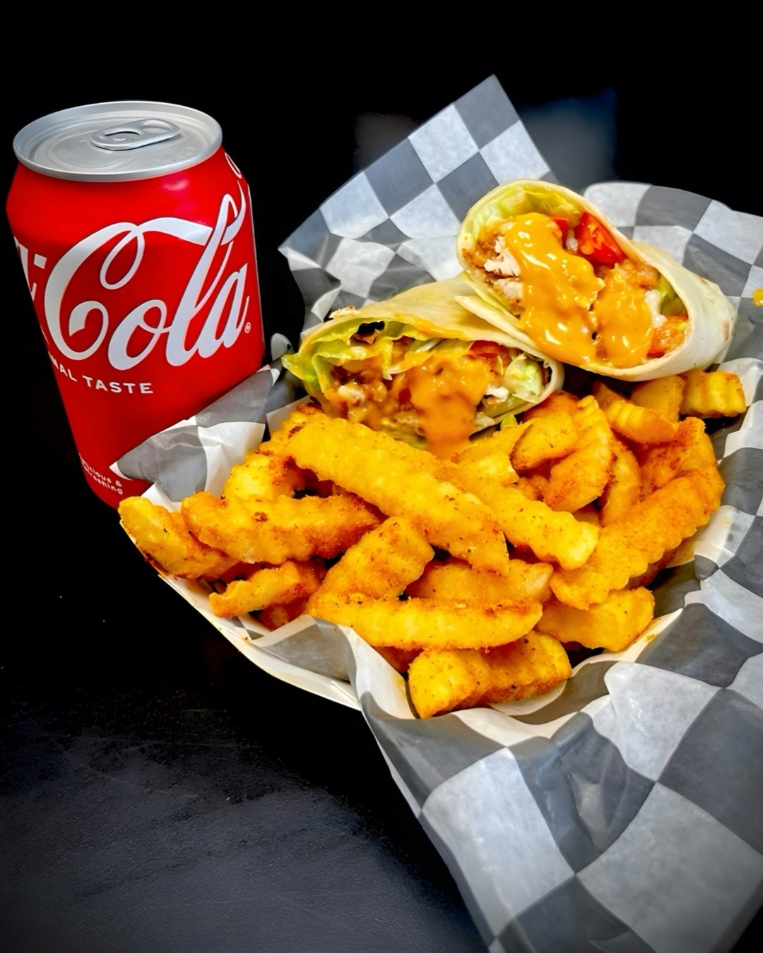 Basket of crinkle-cut fries with two chicken wraps topped with orange sauce, served alongside a can of Coca-Cola.