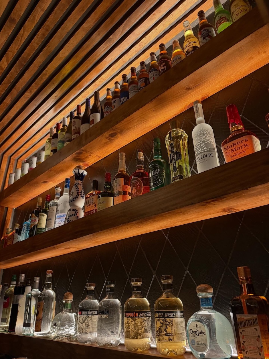 Three wooden shelves mounted on a wall, displaying various bottles of alcoholic beverages including vodka, whiskey, tequila, and beer under warm lighting.