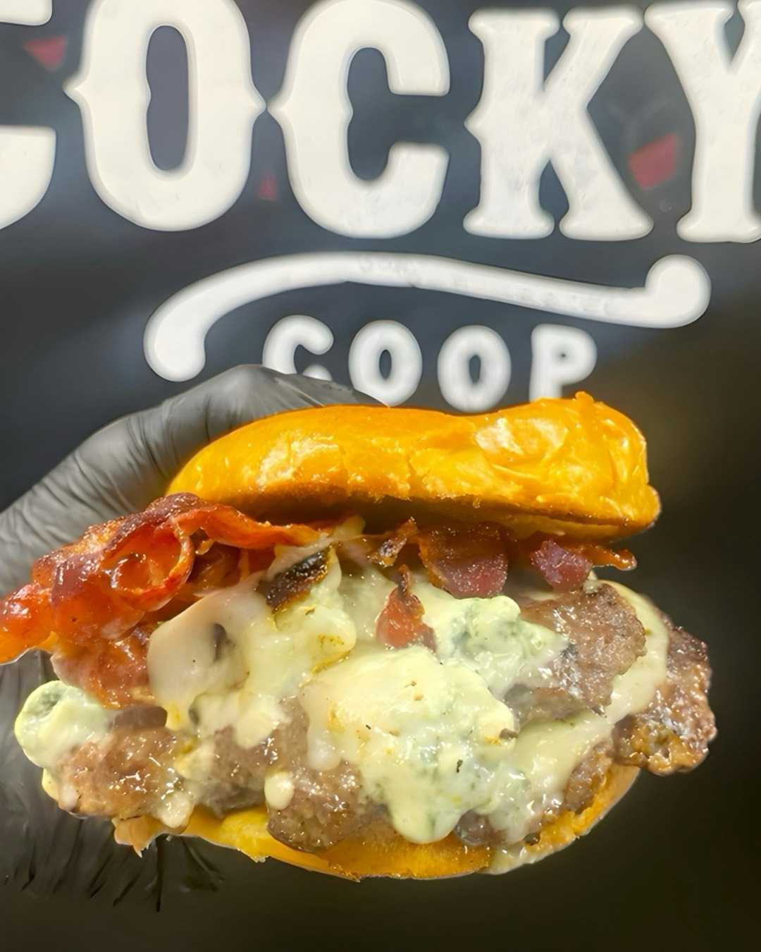 Hand wearing a black glove holding a bacon cheeseburger with melted cheese and a yellow bun against a blurry background with white text.