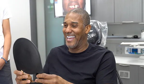 Smiling man in black shirt holding a hand mirror while sitting in a dental chair.