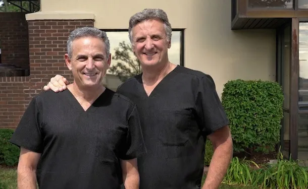 Two men in black scrubs standing side by side outdoors in front of building with greenery.