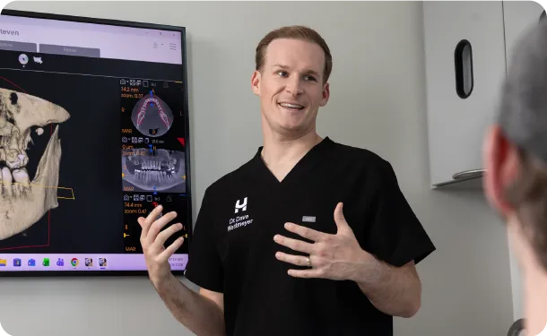 A man in black scrubs explains a dental X-ray displayed on a screen to another person.