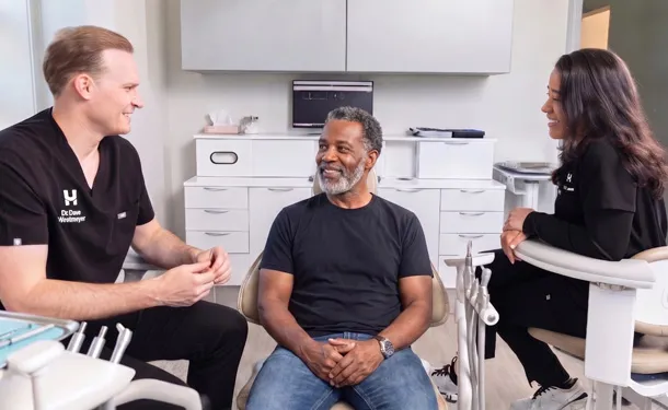 Smiling male patient seated between two healthcare professionals in a bright medical office.