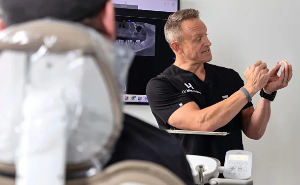 Dentist explaining a dental model to a patient sitting in the dental chair with an X-ray on screen.