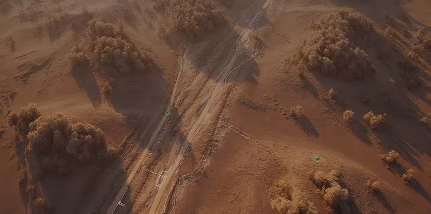 Aerial view of a dirt road winding through dry, brown hills with scattered trees casting long shadows.