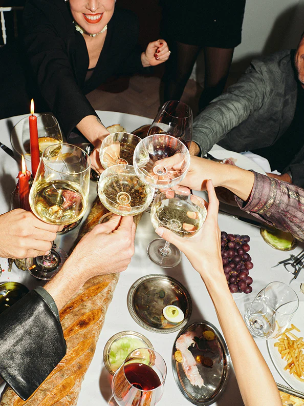 Group of people clinking glasses of white wine over a dinner table with food, candles, and grapes.