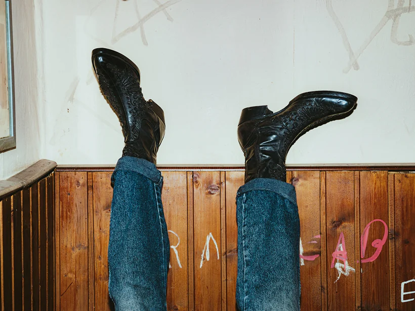 Person wearing cuffed blue jeans and black leather boots with intricate stitching, legs raised against a wooden paneled wall with graffiti.