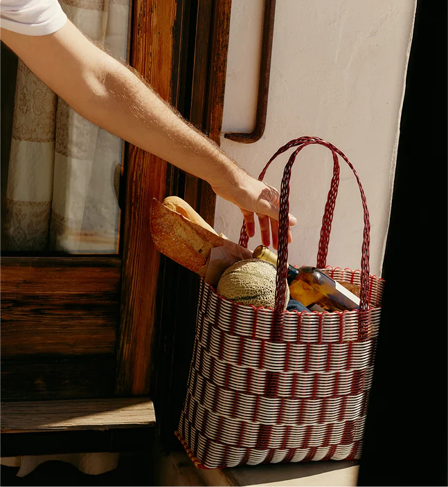 Person placing a woven shopping basket filled with bread, melon, and a bottle at a doorstep.