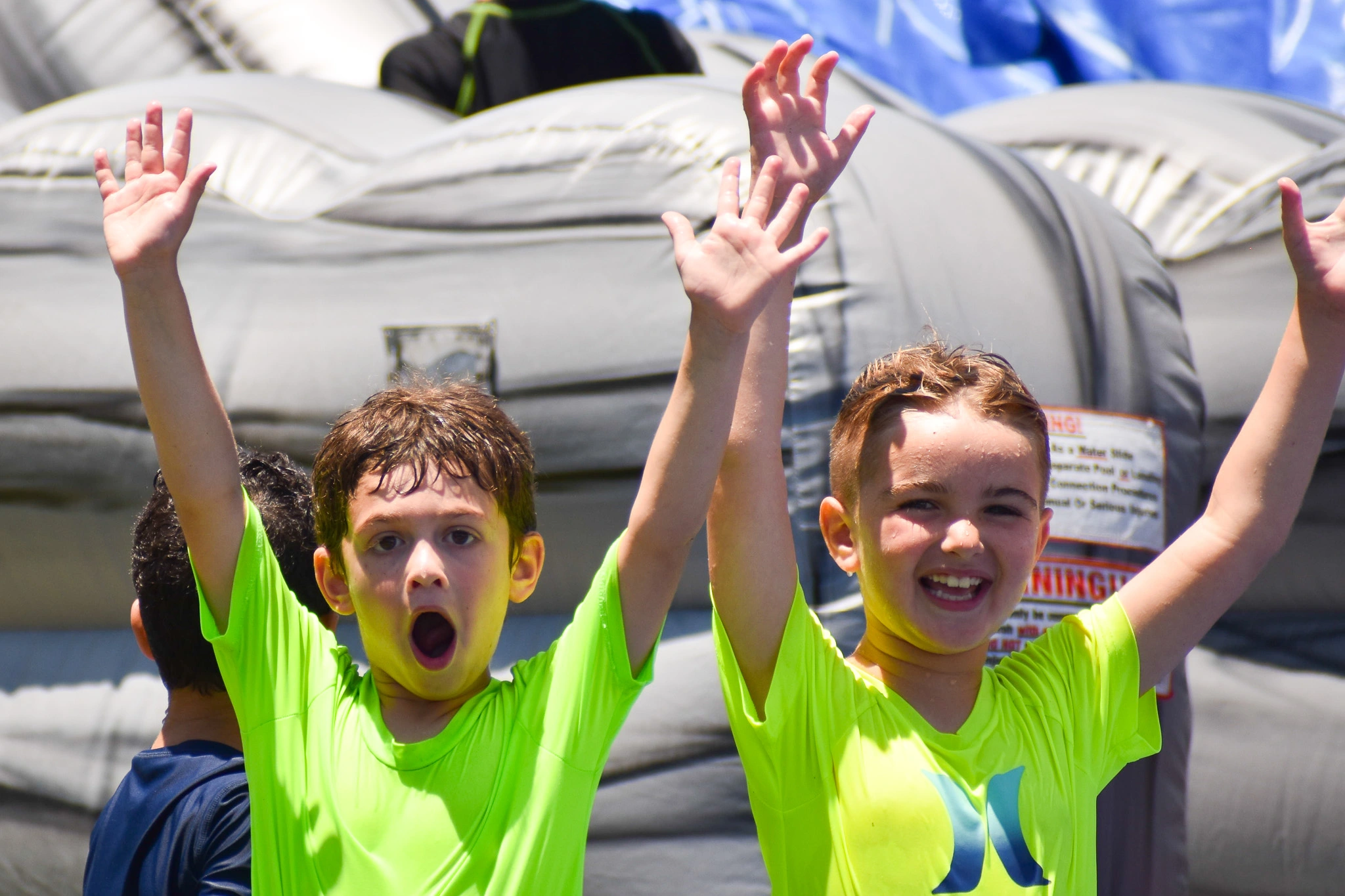 Two children in neon green shirts raising their arms, standing outdoors near an inflatable structure on a sunny day
