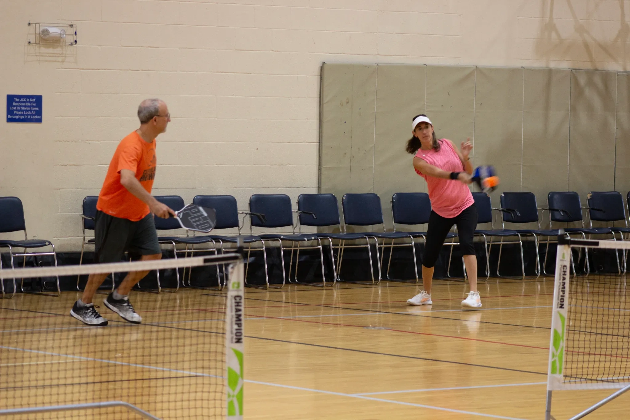 Two people playing pickleball indoors on a wooden court with a net and chairs lined up against the wall