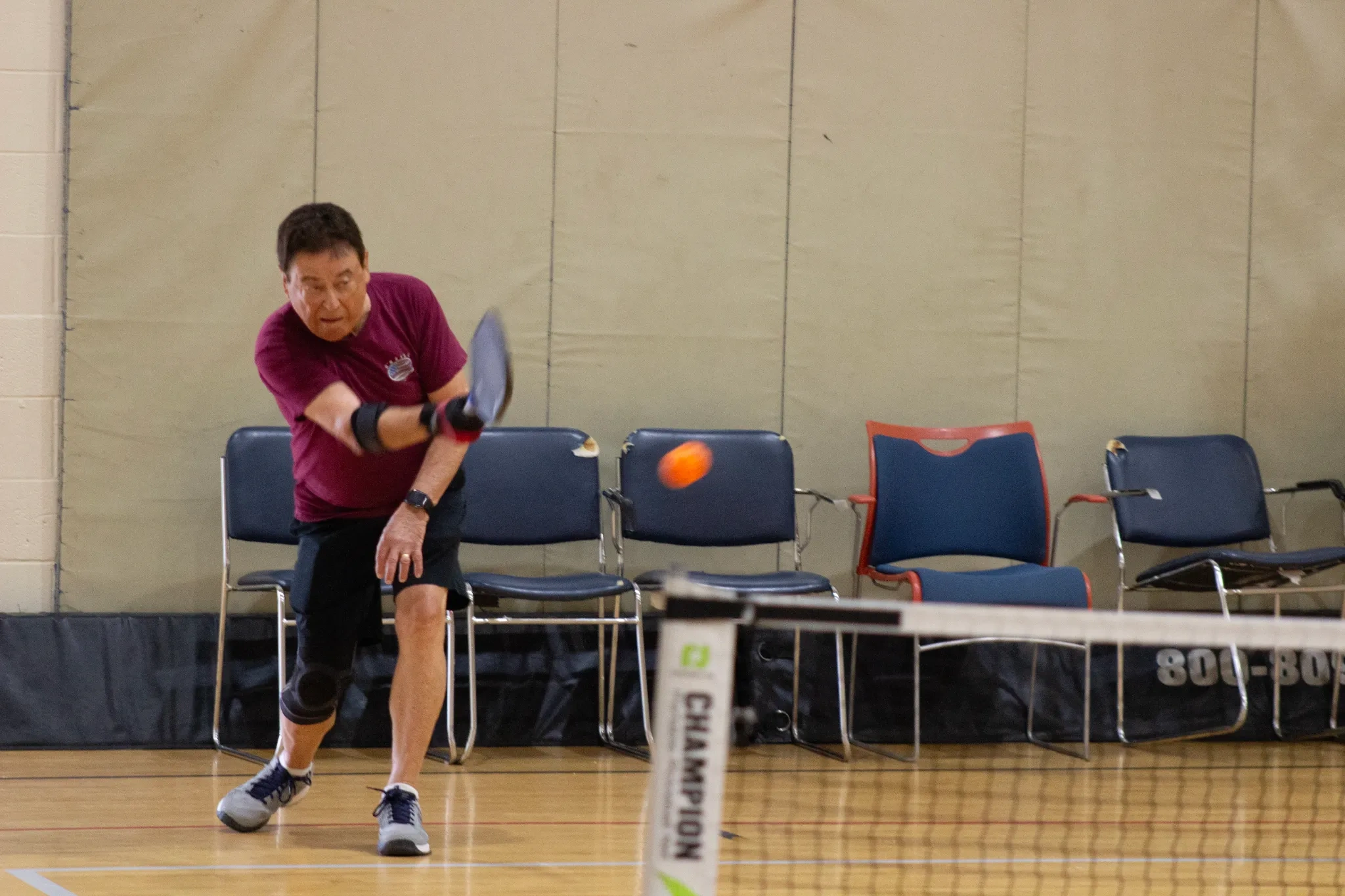 Man playing pickleball indoors, hitting an orange ball over the net, with chairs lined up against the wall in the background
