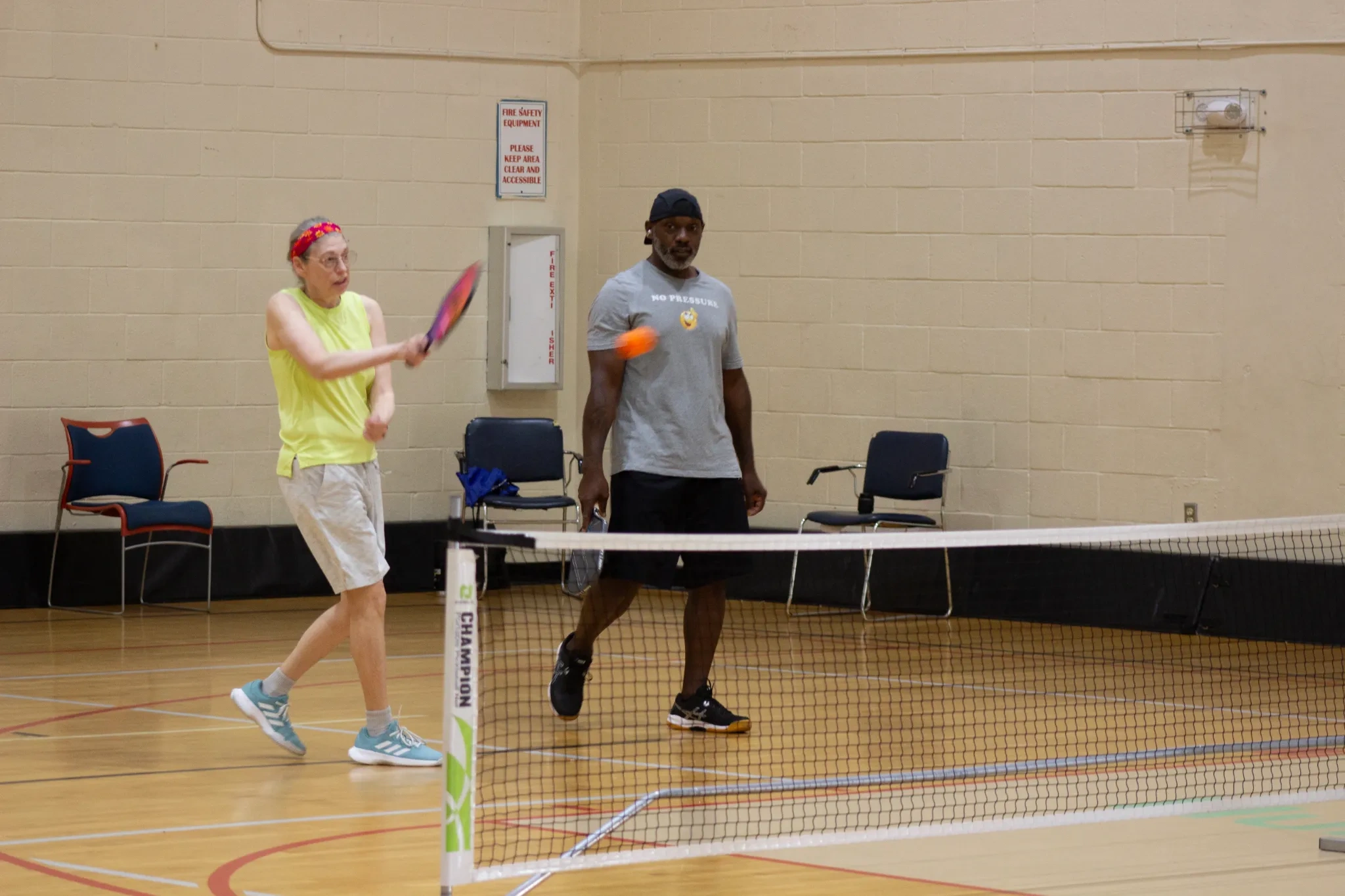 Two people playing pickleball indoors, one hitting the ball over the net while the other watches
