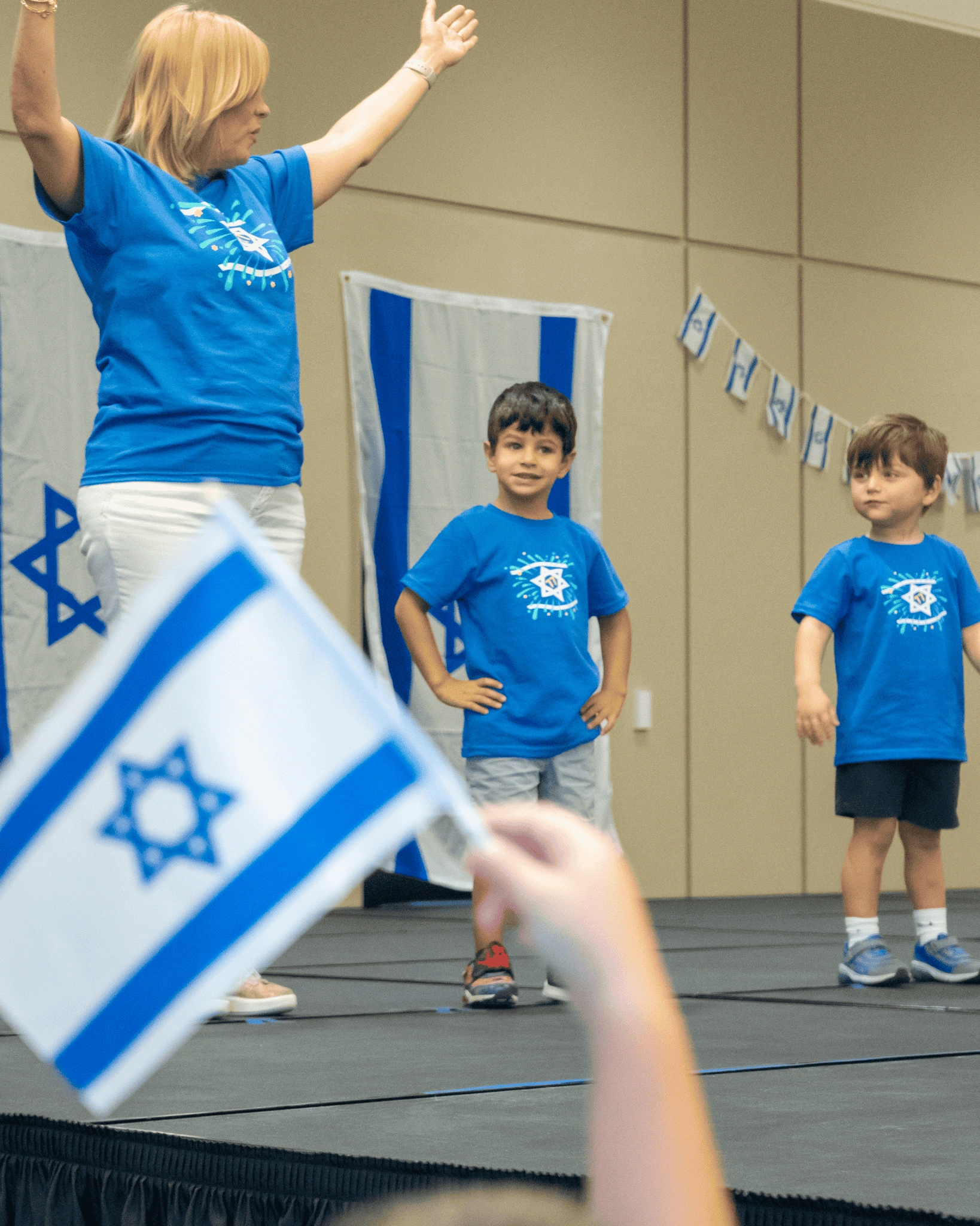 Woman and two children in blue shirts on stage with Israeli flags and decorations, person in foreground waving a flag