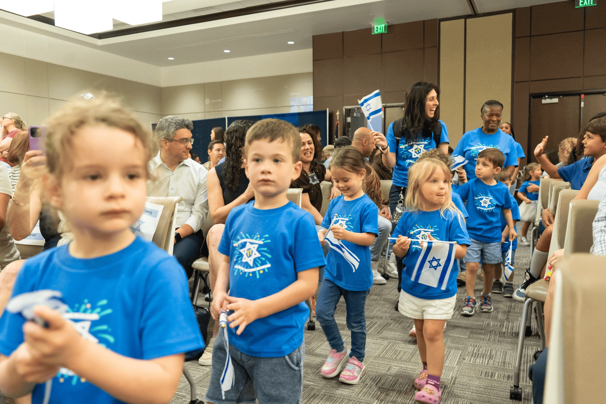 Children in blue shirts holding Israeli flags, walking in a room with seated adults watching an event