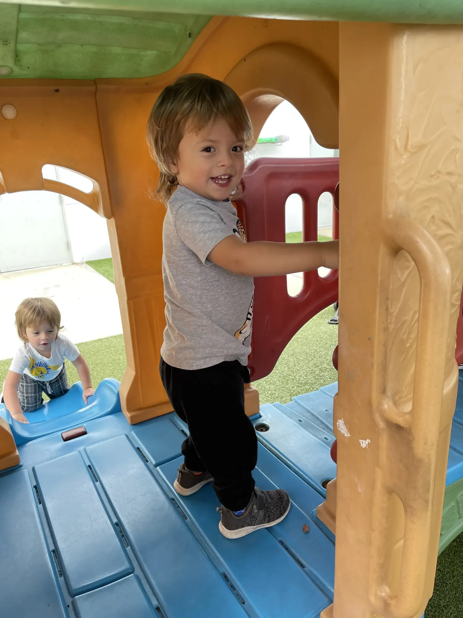Toddler smiling while climbing on a colorful outdoor playground structure with another child nearby