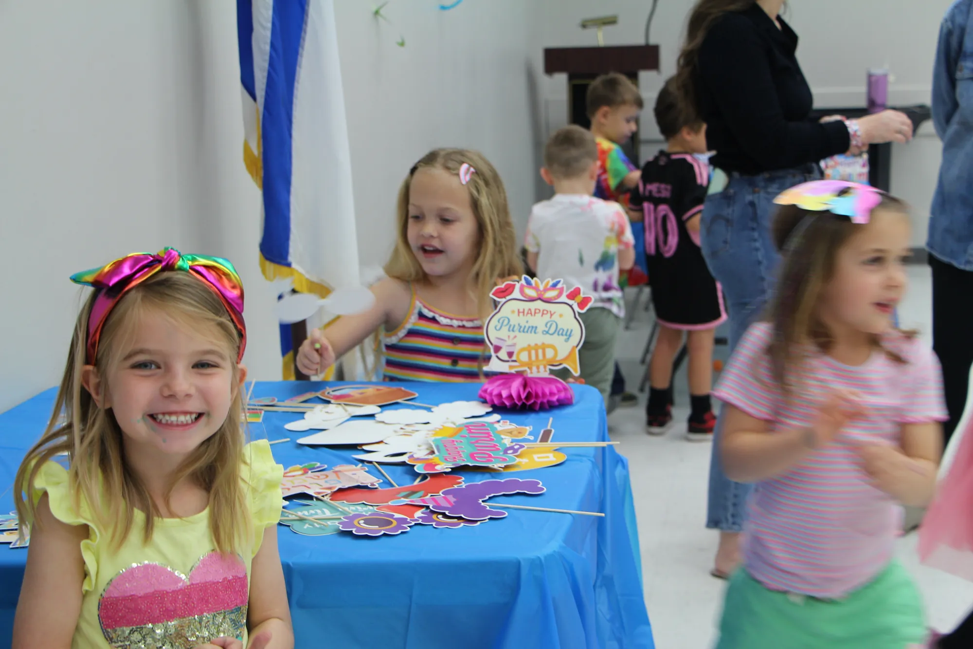 Young preschool girls smiling near a Purim Day craft table decorated with colorful paper props