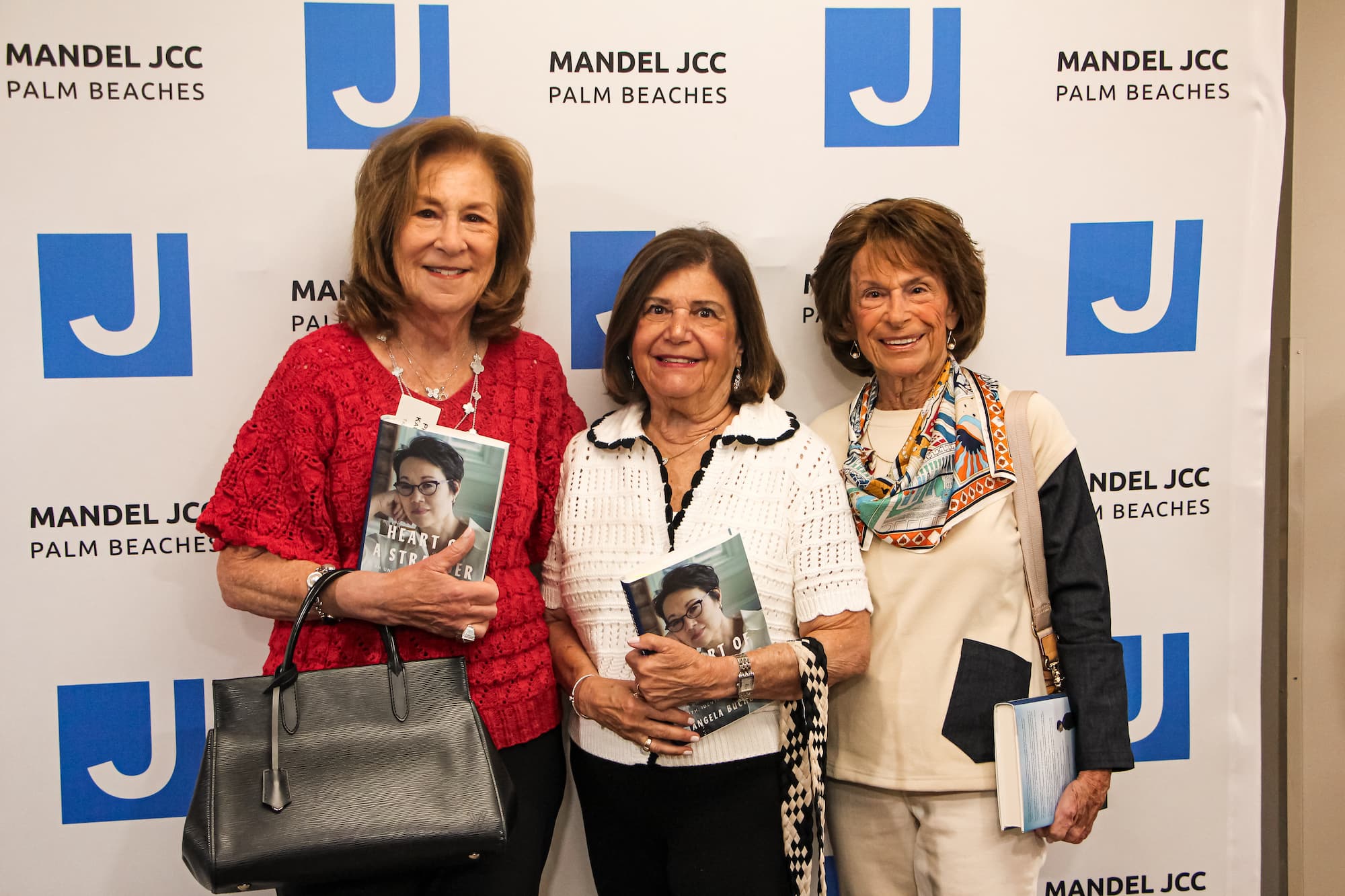 Three elderly women standing in front of a Mandel JCC Palm Beaches backdrop, holding books and smiling at the camera.
