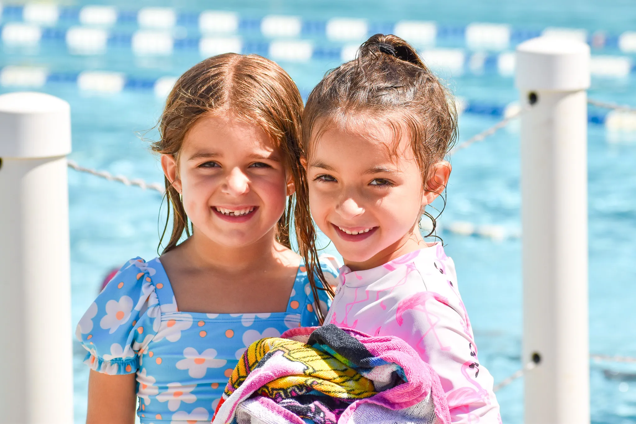 Two smiling young girls in swimsuits standing by a pool, one holding colorful towels.