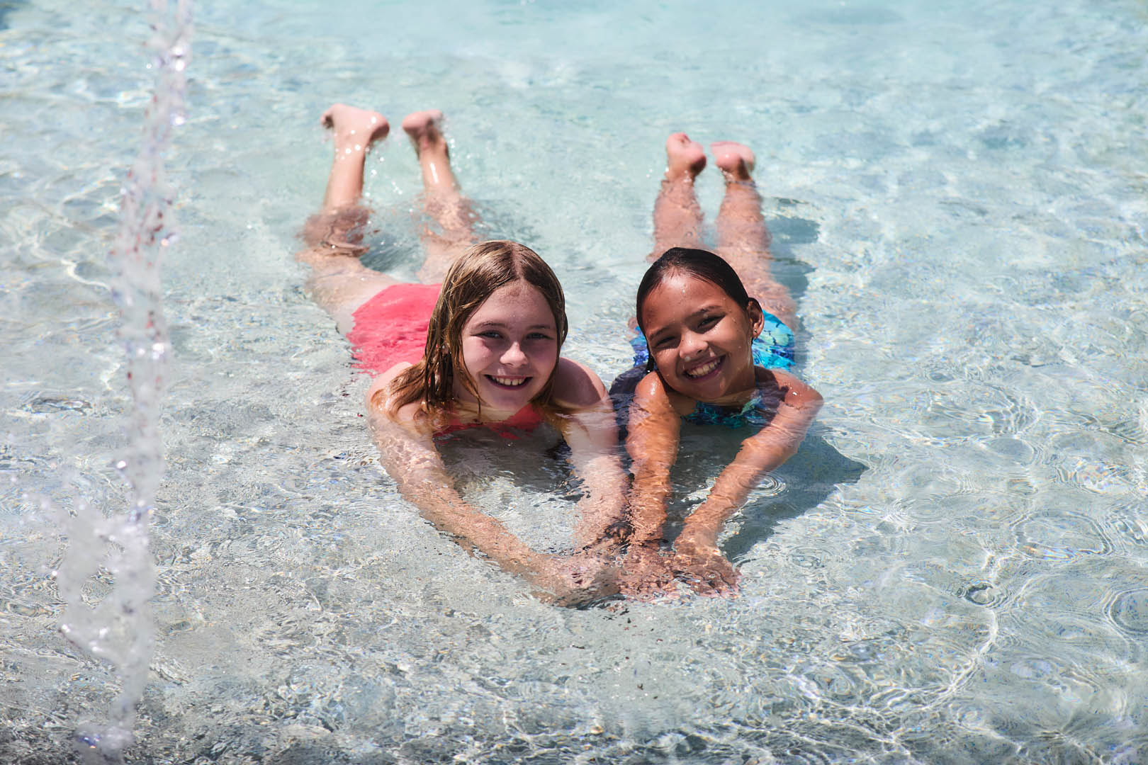Two smiling girls lying on their stomachs in shallow clear pool water holding hands.