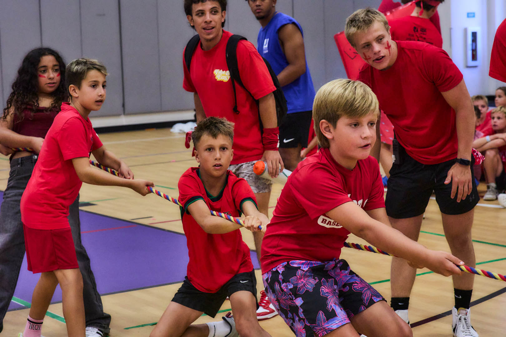 Children and young adults in red shirts playing tug-of-war indoors on a gym floor, with some coaches or older participants observing.