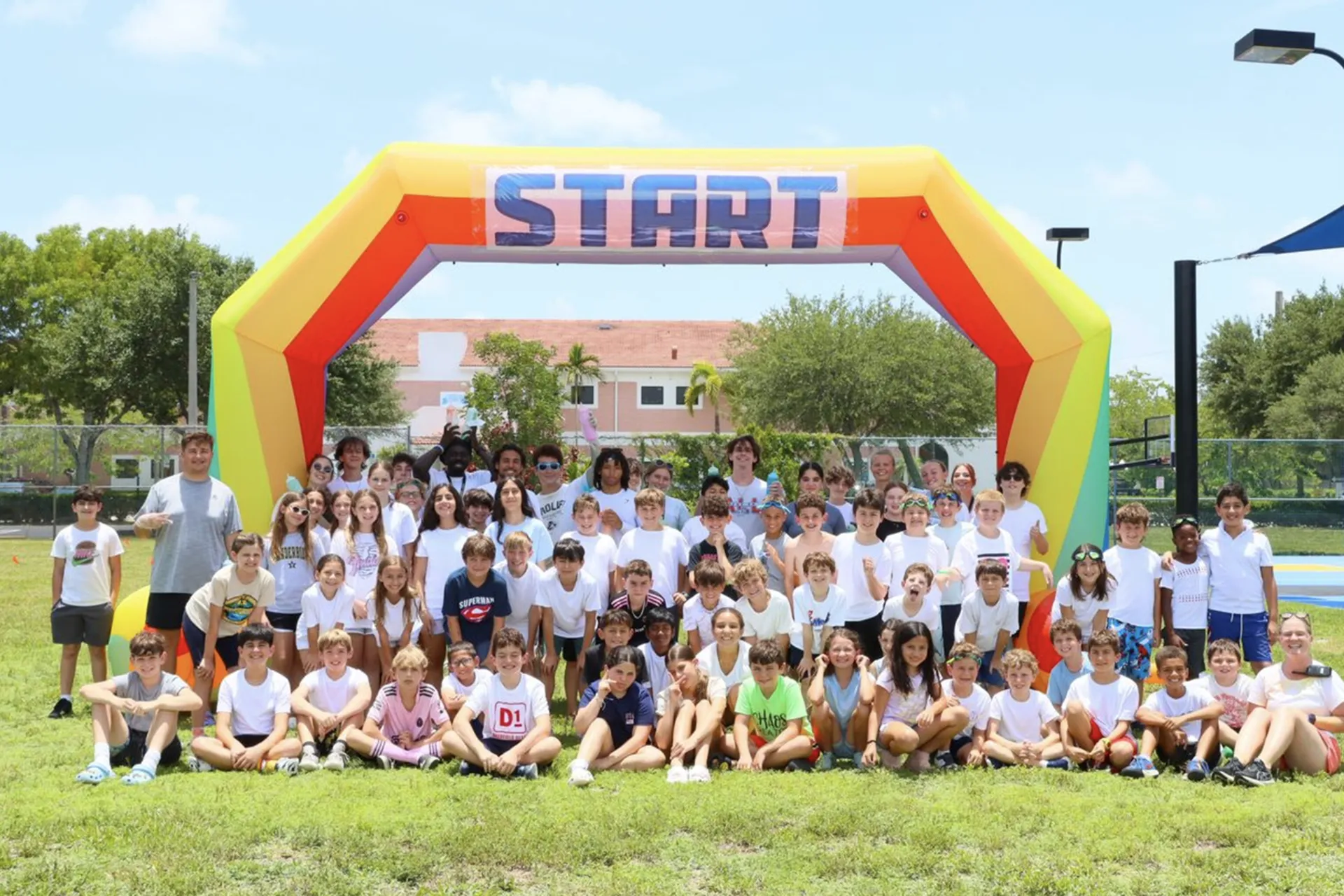 Large group of children and adults posing under a colorful inflatable arch with the word START on a sunny day outdoors.