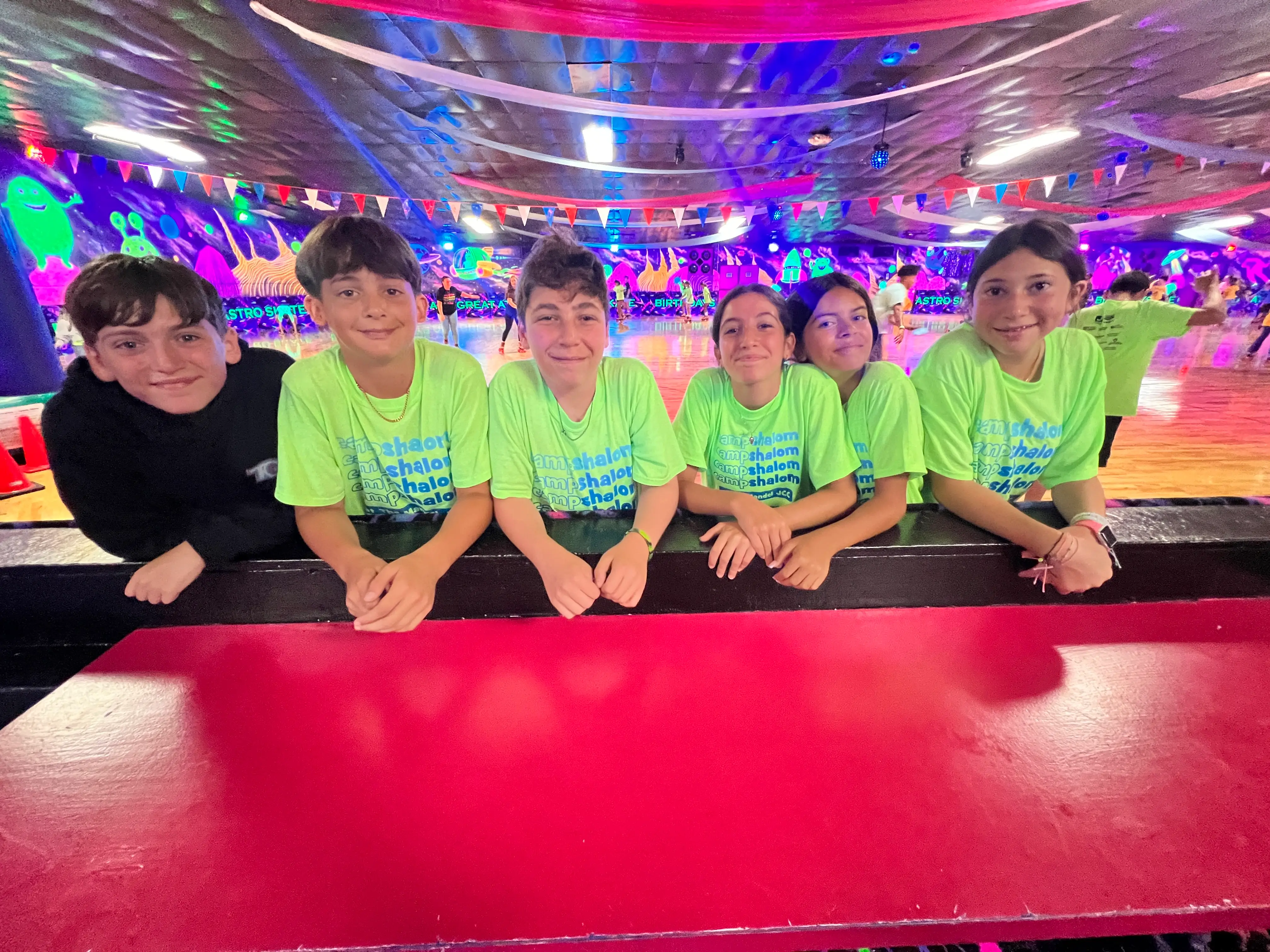 Six children smiling and leaning on a railing at a roller skating rink with neon-lit walls and bunting overhead.