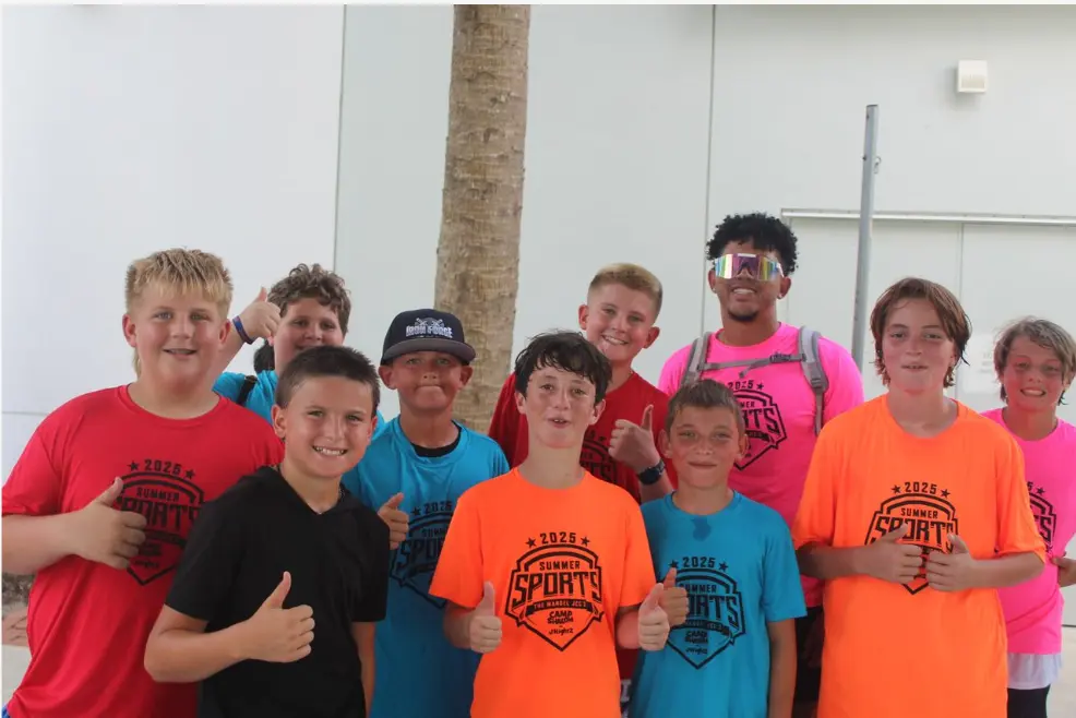 Group of smiling children and a coach wearing colorful 2025 Summer Sports camp t-shirts posing outdoors with thumbs up.