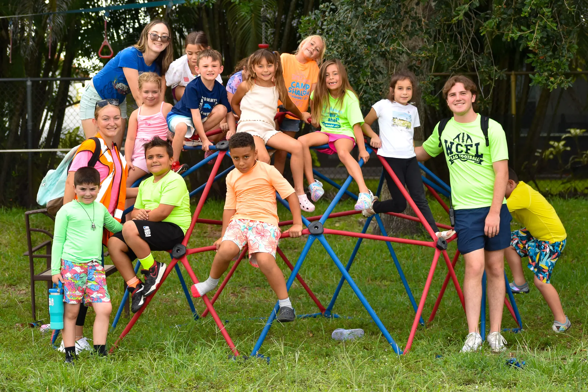 Group of children and two adults smiling and playing on a red and blue jungle gym in an outdoor grassy area.