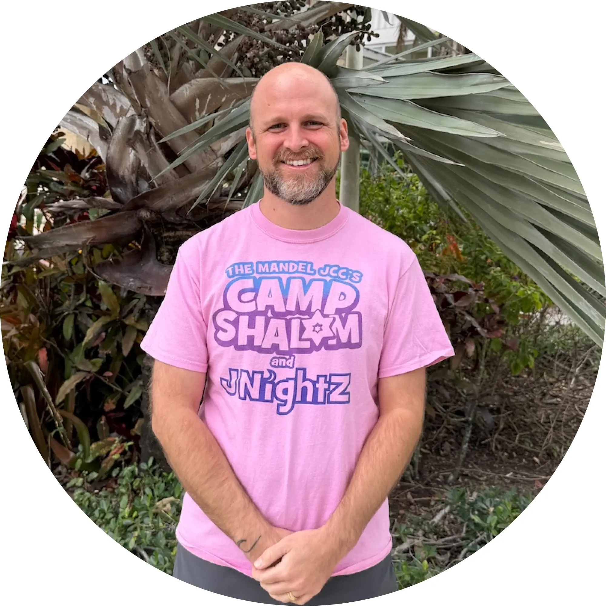 Smiling bald man with beard wearing a pink Camp Shalom and JNightz t-shirt standing outdoors in front of tropical plants.