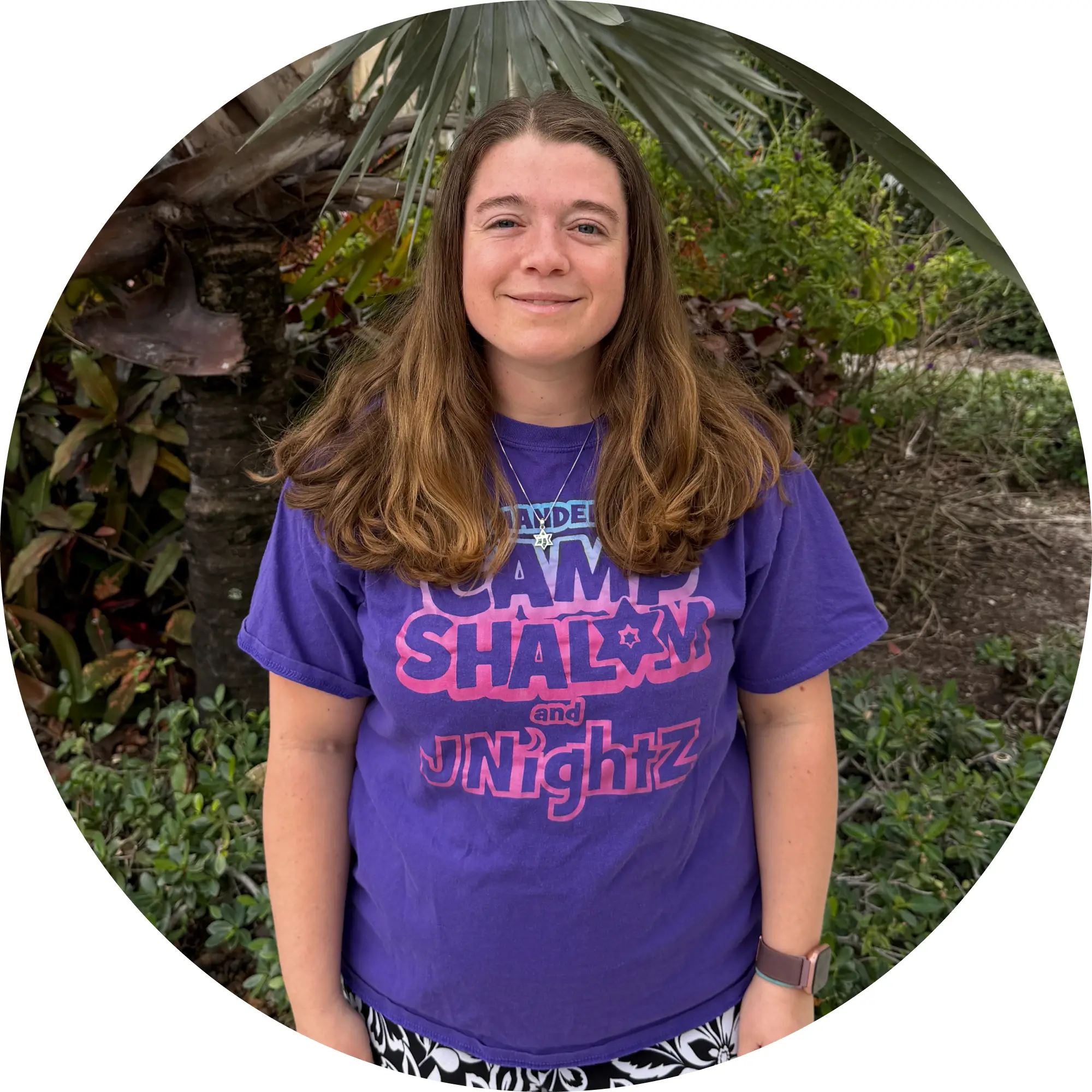 Young woman with long brown hair wearing a purple Camp Shalom and JNIGHTZ t-shirt, standing outdoors in front of greenery and a palm tree.