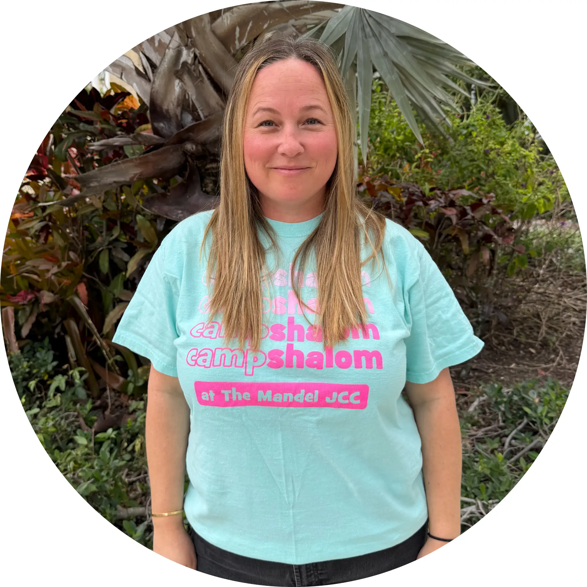 Woman with long blonde hair smiling, wearing a light blue T-shirt with pink text that says 'camp shalom at The Mandel JCC,' standing outdoors with green plants in the background.