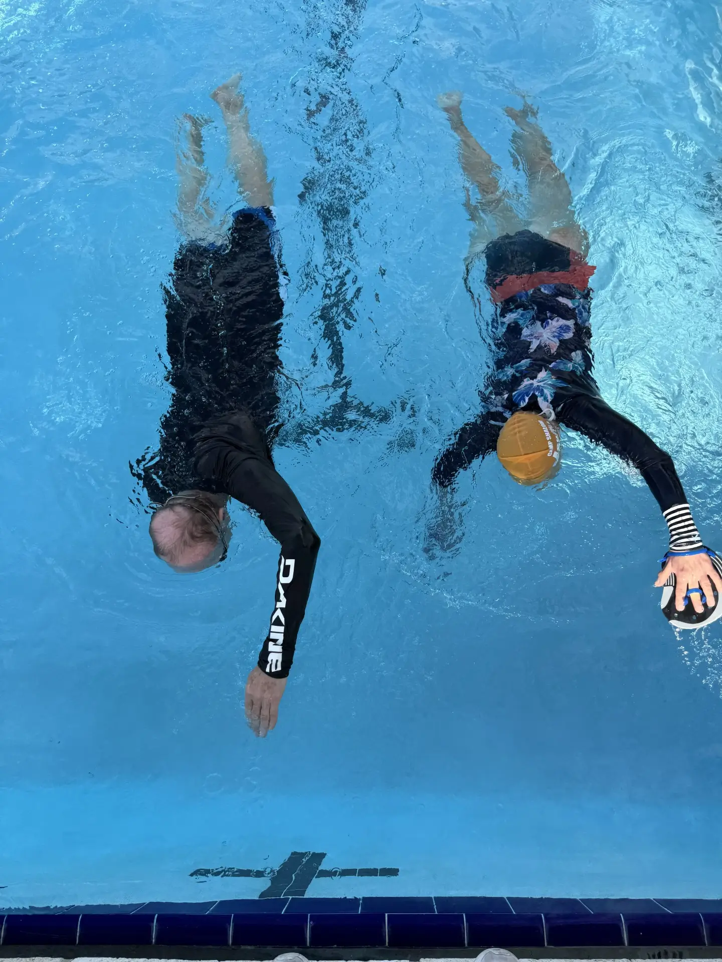 Two people swimming underwater side by side in a pool, one holding a water polo ball.