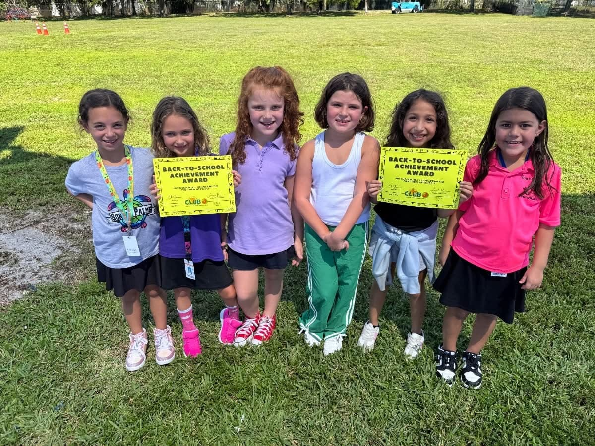 Group of six smiling young girls standing on grass outdoors, two holding yellow back-to-school achievement award certificates.