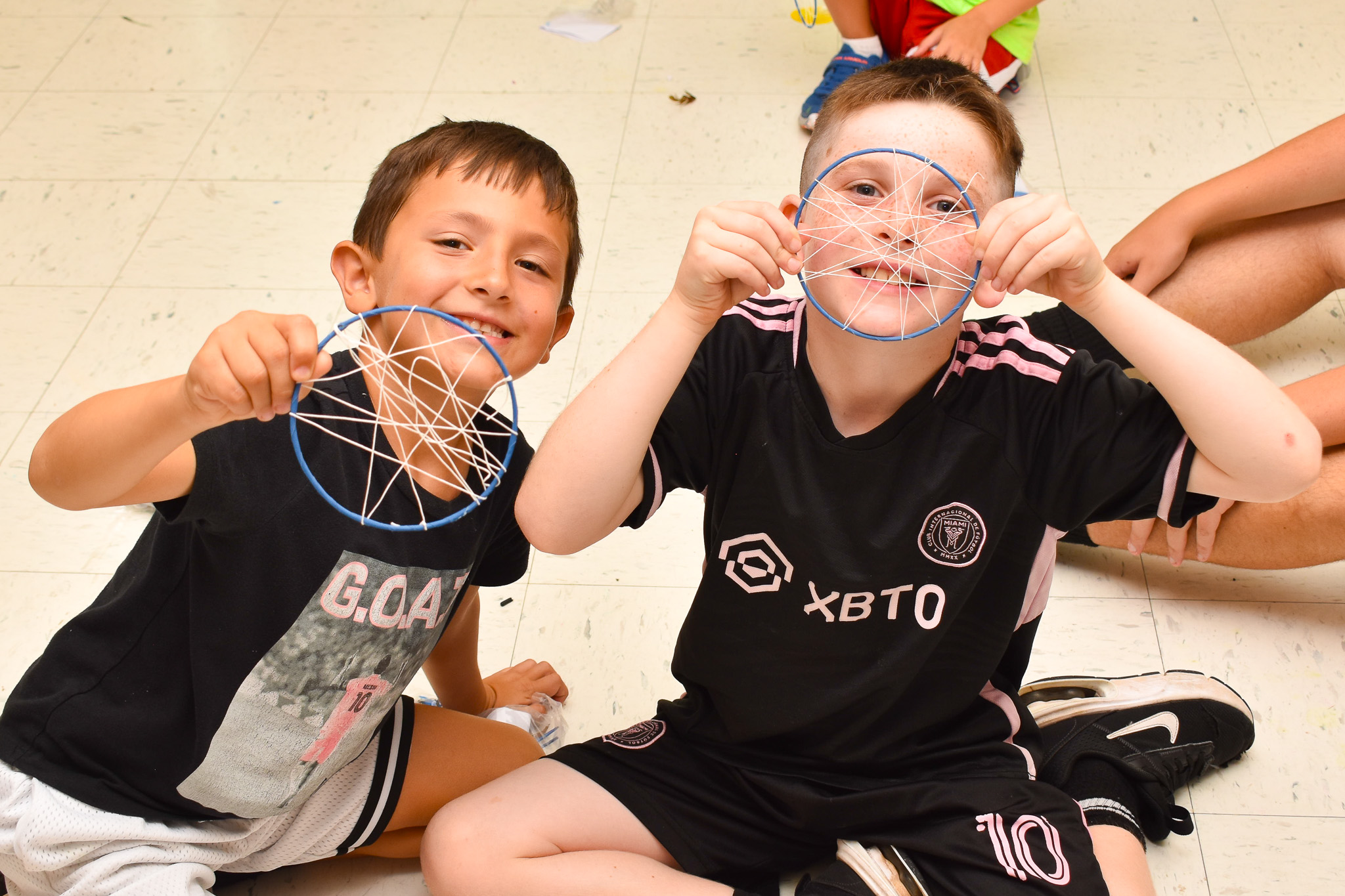 Two boys sitting on a tiled floor smiling and holding up blue circular crafts with white string webbing.