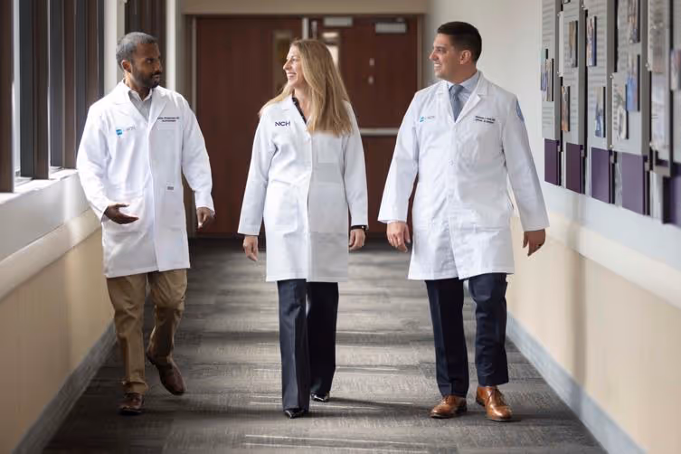 Three medical professionals in white coats walking and talking in a hospital corridor.