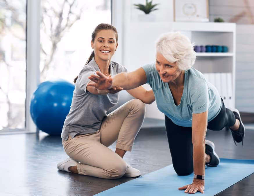 Young female trainer guiding an older woman doing a balance exercise on a yoga mat in a fitness studio.